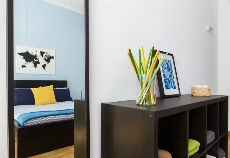 Camera da letto moderna visibile in gran parte attraverso uno specchio alto, con copriletto a tema, cuscini blu e un cuscino giallo; davanti un mobile a cubi con oggetti decorativi crea un gradevole angolo d'ingresso.