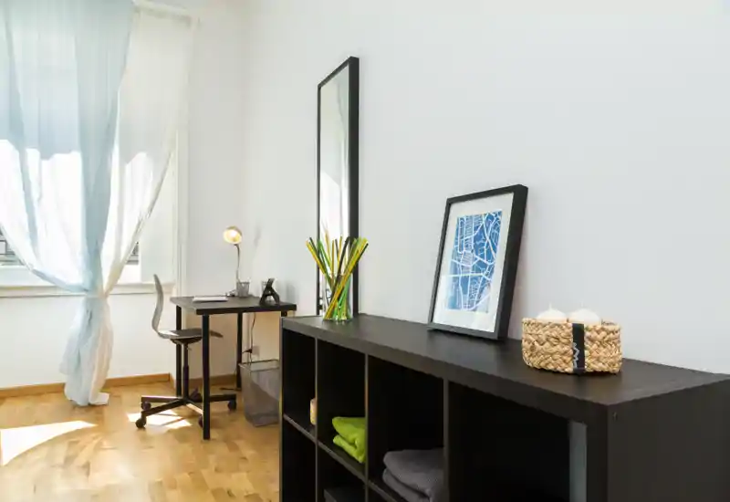 Zona giorno luminosa e minimal con una scrivania compatta, mobile a cubi e luce naturale da un’ampia finestra — ideale anche come angolo studio.
