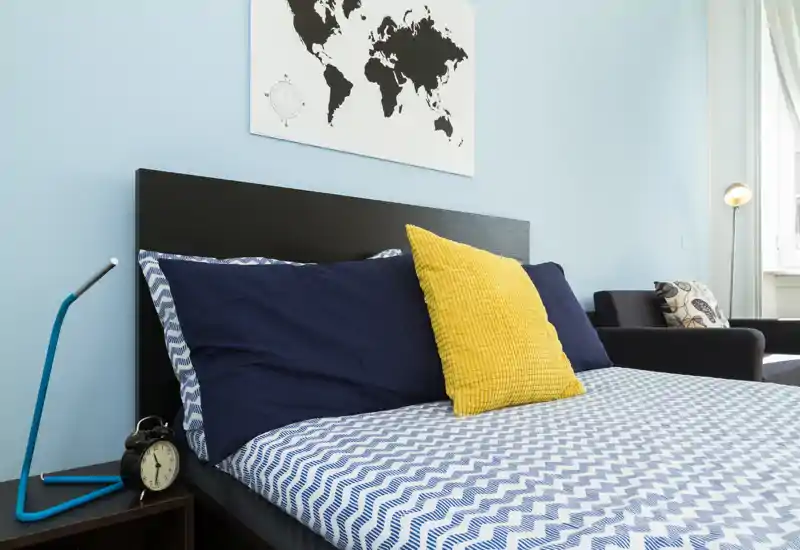Camera da letto moderna e luminosa con lenzuola a motivo blu e bianco, cuscini blu navy e un cuscino giallo a contrasto; quadro mappamondo sopra la testiera e un angolo salotto vicino alla finestra.