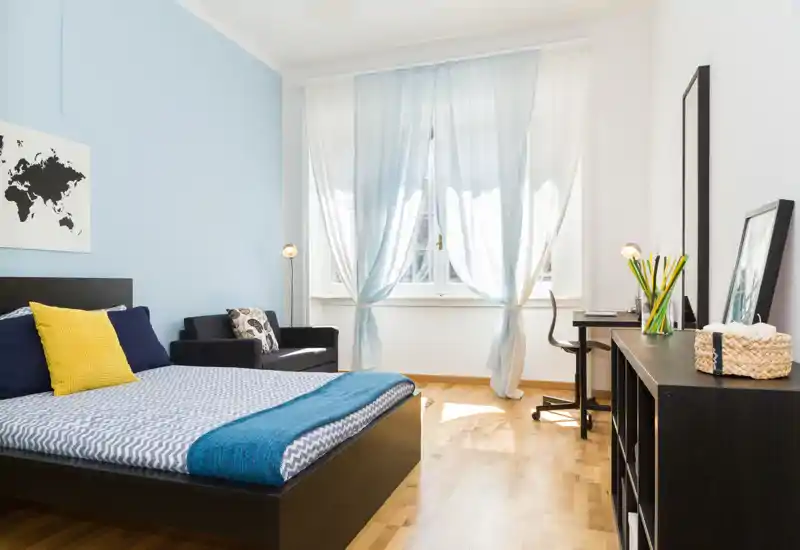Camera da letto luminosa e moderna con letto matrimoniale, accenti azzurri e luce naturale da una grande finestra; comprende una piccola zona salotto e una scrivania.