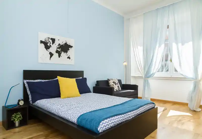 Camera da letto luminosa e di stile con letto matrimoniale, cuscini blu e gialli, una poltrona e grandi finestre che lasciano entrare molta luce naturale.