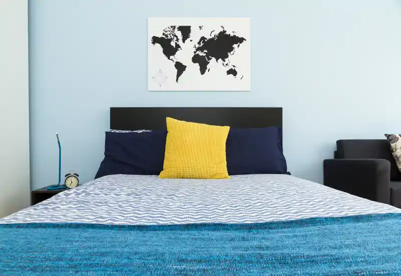 Camera da letto luminosa e moderna con un letto matrimoniale ben rifatto, cuscini blu navy e un cuscino giallo a contrasto, con una stampa mappa del mondo sopra la testata.