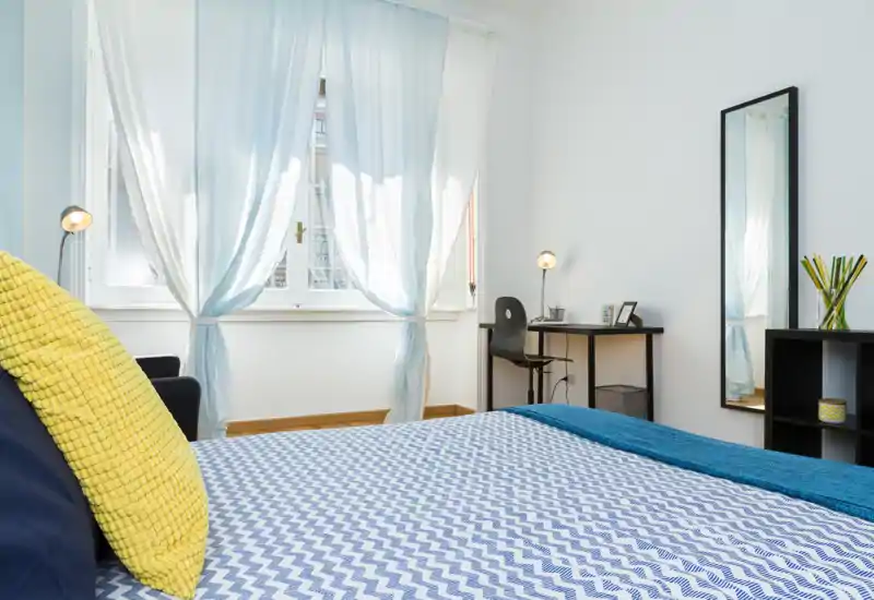 Camera da letto luminosa e ben arredata con un grande letto in primo piano, biancheria blu e bianca, un cuscino giallo come accento e una piccola scrivania vicino alla finestra.