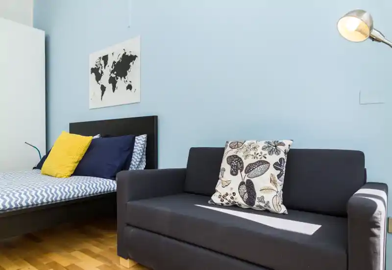 Camera da letto luminosa e ordinata con letto ben visibile e divano scuro in primo piano; pareti azzurre e cuscini decorativi creano un ambiente moderno e rilassante.