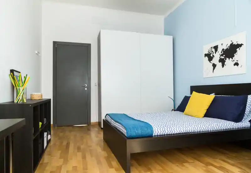 Camera da letto moderna e luminosa con letto matrimoniale, cuscini colorati e un armadio. Linee pulite, pavimento in legno e una semplice stampa murale creano un ambiente accogliente.