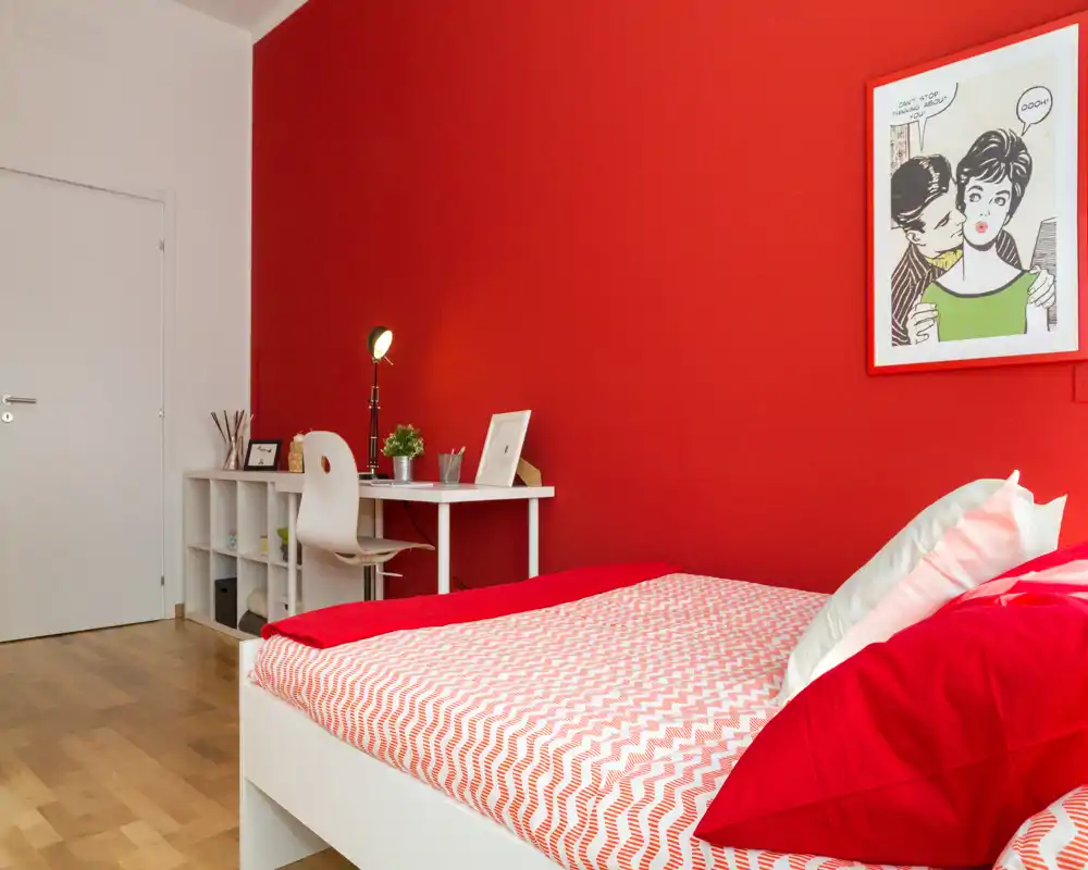 Dormitorio moderno y luminoso con pared roja y una cama bien hecha en primer plano; un pequeño escritorio y estantería junto a la pared crean un espacio funcional y con estilo.