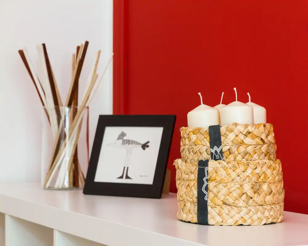 Detalle de sala con cestas tejidas decorativas, velas, cuadro y jarrón de cañas frente a una pared roja — un rincón acogedor ideal para fotos de anuncio.