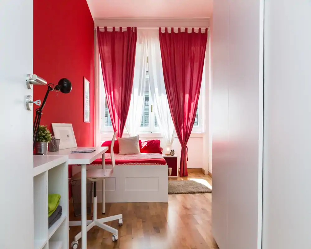 Dormitorio luminoso y con estilo con una cama tipo sofá blanco bajo una gran ventana con cortinas rojas; escritorio y silla a un lado, textiles acogedores y suelo de madera cálida.
