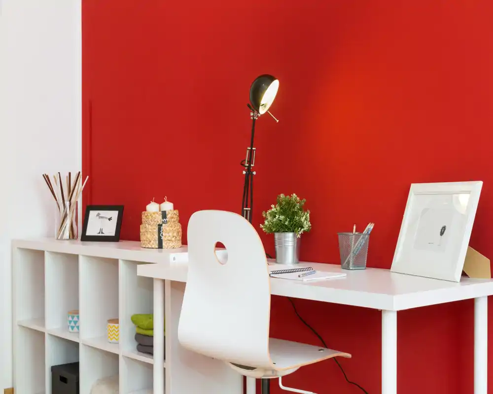Atractivo rincón de oficina en casa luminoso con escritorio blanco, silla moderna, lámpara de trabajo y detalles decorativos frente a una pared roja.