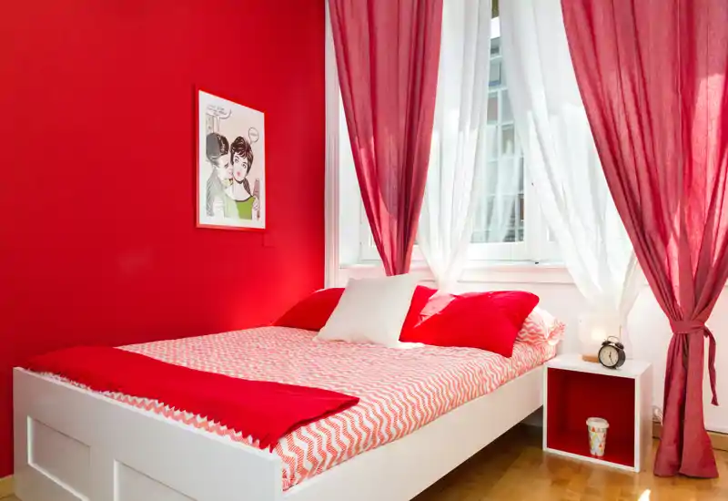 Dormitorio moderno y luminoso con cama baja blanca, textiles rojos y una gran ventana que aporta abundante luz natural: acogedor y con estilo para un anuncio.
