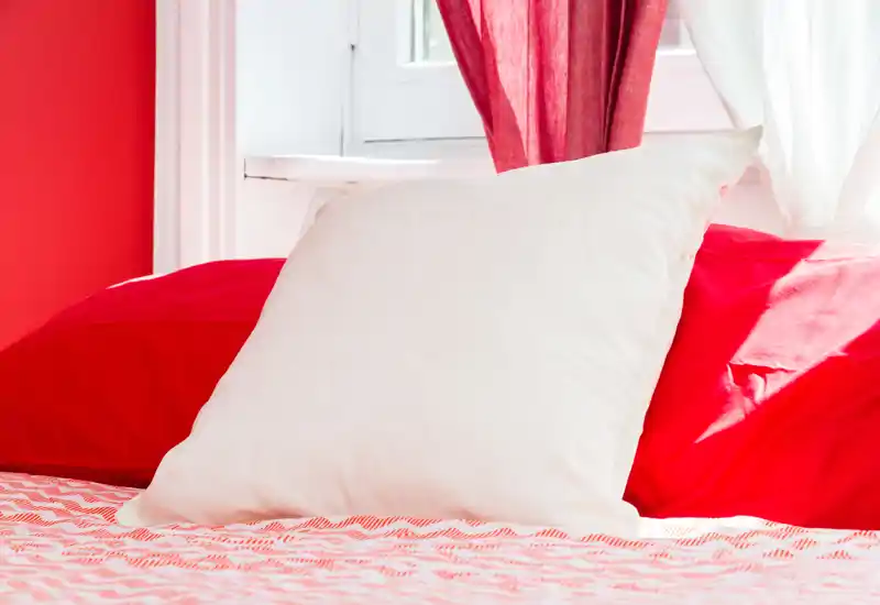 Primer plano de un dormitorio luminoso y acogedor con almohadas y edredón rojo frente a una ventana soleada, ideal para destacar la cama en un anuncio.