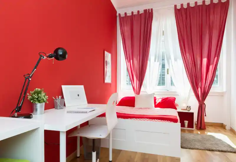 Dormitorio luminoso y acogedor con cama individual junto a una ventana grande, cortinas rojas y muebles blancos; incluye un escritorio pequeño para trabajar.