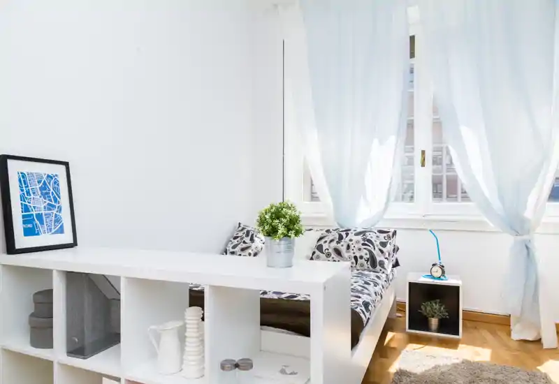 Camera da letto luminosa e minimal con letto confortevole, cuscini decorativi e un piccolo comodino accanto alla finestra che lascia entrare molta luce naturale.