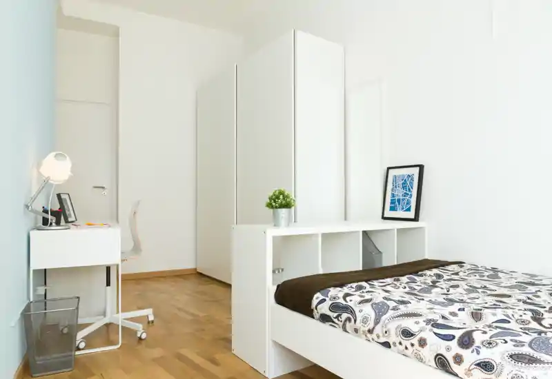 Camera da letto luminosa e minimalista con letto singolo, mensole compatte a piedi del letto, una piccola scrivania e armadio — ambiente pulito e ben illuminato ideale per una persona.