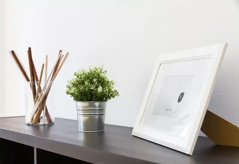 Mensola decorativa minimal con una piccola pianta in vaso, una cornice fotografica e un vaso di vetro con stecche di bambù, ripresa in un primo piano pulito e ben illuminato.