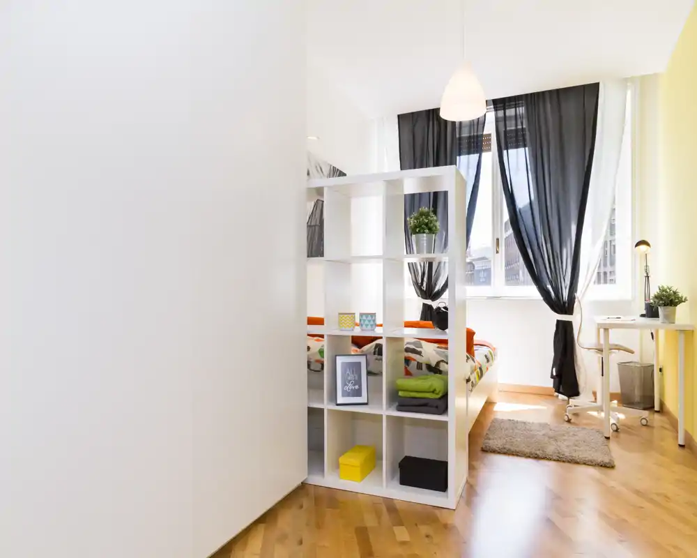 Chambre claire et moderne avec lit visible derrière une étagère ouverte en cubes, grande fenêtre avec rideaux foncés, petit bureau et sol en bois — idéale pour les annonces mettant en avant la luminosité et le design fonctionnel.