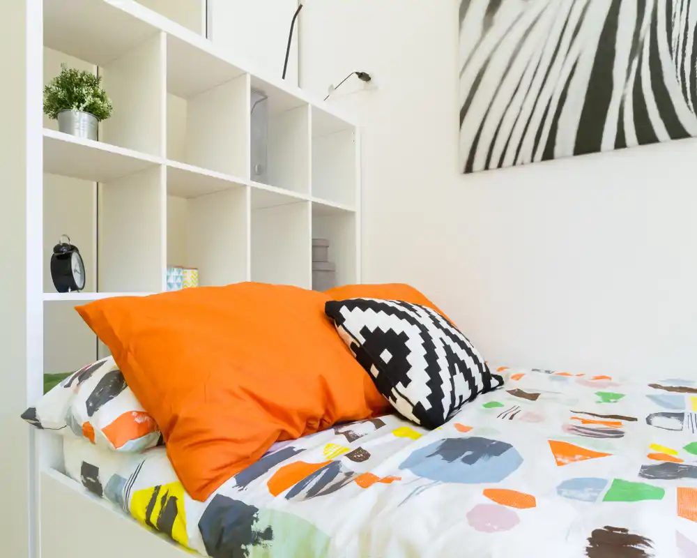 Photo lumineuse et moderne d'une chambre, centrée sur un lit coloré avec une couette à motifs et des coussins décoratifs, devant une étagère blanche et une œuvre murale.