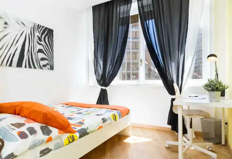 Camera da letto luminosa e moderna con letto singolo e biancheria colorata, ampie finestre con tende nere semitrasparenti, piccolo scrittoio e sedia — perfetta per uno studente o una persona sola.