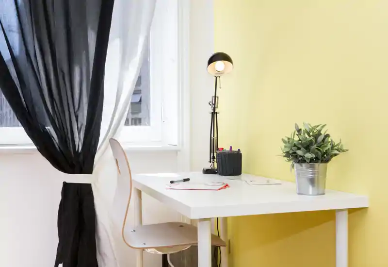 Angolo scrittoio luminoso e ordinato con una sedia semplice, lampada e una pianta in vaso su una parete gialla calda — ideale per un compatto angolo studio o smart working.