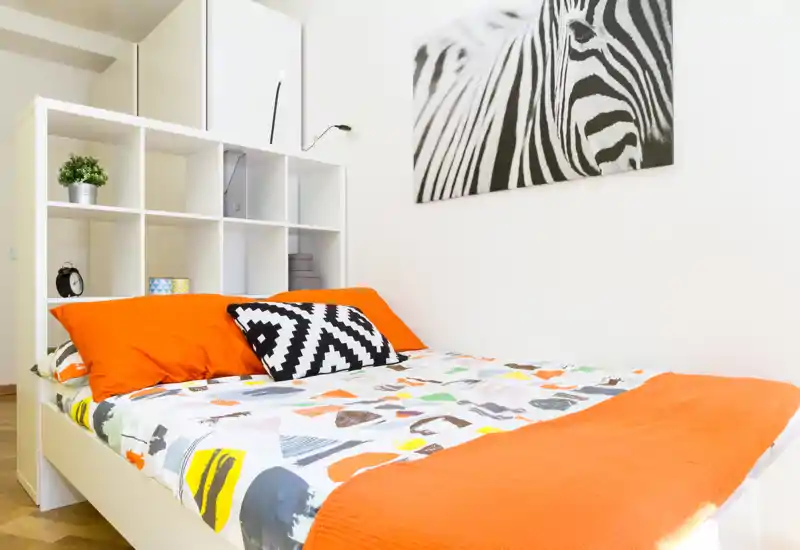Camera da letto moderna e luminosa con copripiumino colorato, accenti arancioni vivaci e una stampa zebracava alle pareti; una libreria a cubi bianca dietro il letto offre spazio e stile.