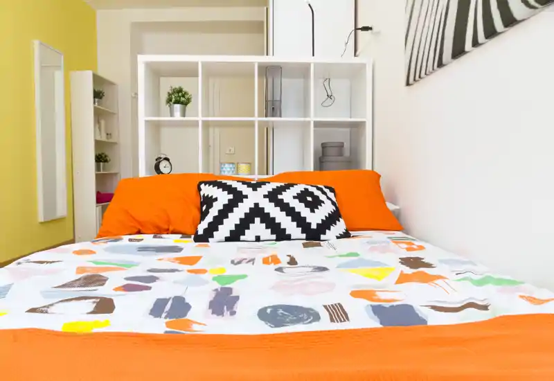 Camera da letto luminosa e ben allestita con una coperta colorata e un cuscino dal motivo deciso; un mobile a cubi bianco dietro il letto offre spazio e profondità visiva.