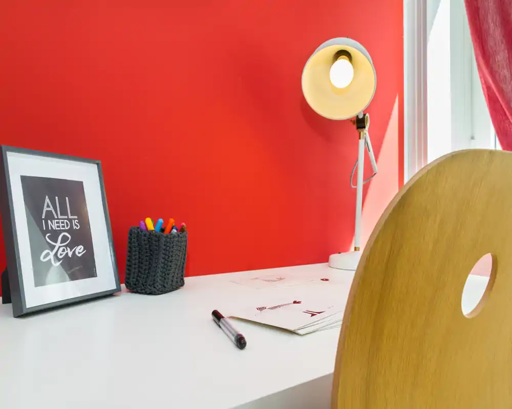 Rincón de trabajo moderno y luminoso con superficie blanca, silla de madera, lámpara de escritorio y bolígrafos de colores frente a una pared roja intensa — ideal como despacho o zona de estudio.
