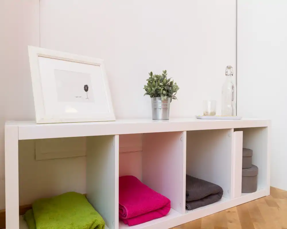 Esquina de salón luminosa y minimalista con estantería abierta, mantas de colores dobladas y una pequeña planta: un espacio auxiliar ordenado y acogedor.