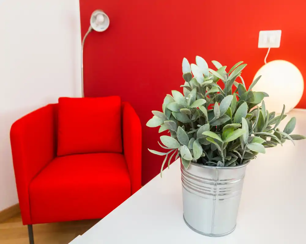 Rincón de salón luminoso y con estilo con un sillón rojo intenso, planta decorativa en maceta metálica y luz cálida; ideal para un rincón de lectura acogedor.