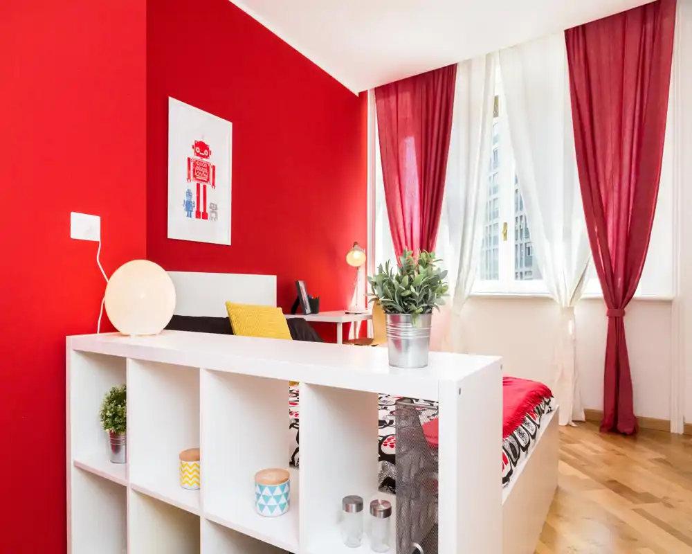 Dormitorio moderno y luminoso con pared roja de acento, cama blanca y estantería divisoria en primer plano; grandes ventanas con cortinas rojas y blancas que proporcionan mucha luz natural, decorado con plantas y cojines de colores.