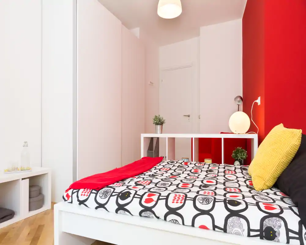 Dormitorio moderno y luminoso con colcha estampada, cojines de colores y muebles blancos; la pared roja aporta carácter y el ambiente resulta fresco y acogedor.