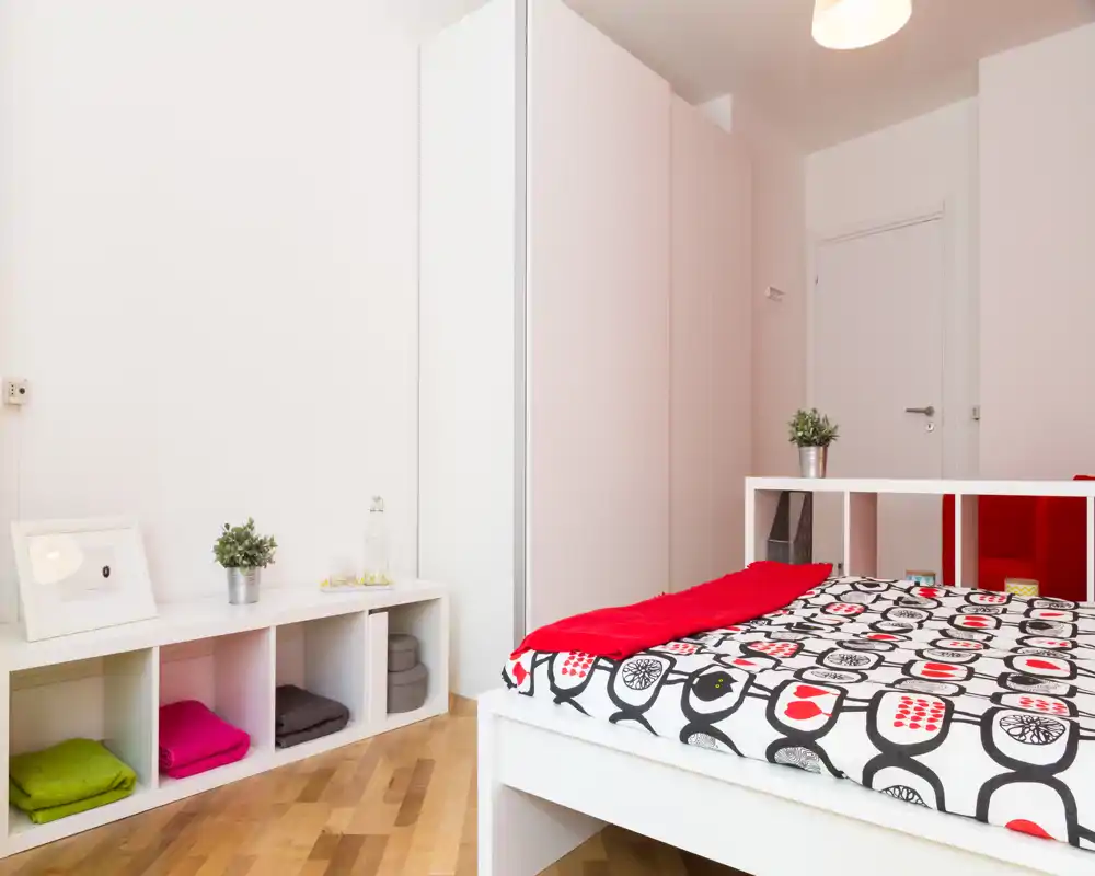 Dormitorio moderno y luminoso con colcha estampada y manta roja sobre una cama de estructura blanca, estantería minimalista y armario; espacio ordenado y bien iluminado con pequeñas plantas decorativas.