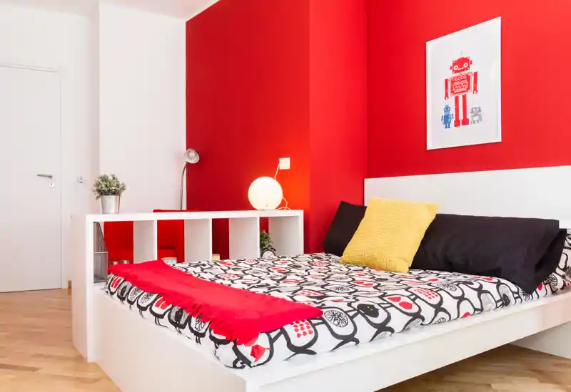 Chambre moderne et lumineuse avec un lit double complet, un mur rouge vif et une literie à motifs, offrant une ambiance chaleureuse et stylée.