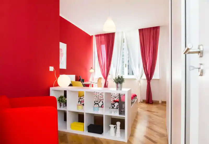 Chambre lumineuse et tendance avec lit à motif, mur d'accent rouge et voilages laissant entrer la lumière naturelle. Une étagère moderne sépare l'espace nuit du coin salon, créant une atmosphère chaleureuse et bien mise en scène.