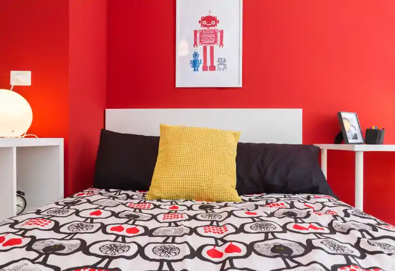 Chambre lumineuse et stylée avec couette à motif, coussin jaune et murs rouge vifs. Le lit est au centre, éclairé par une lampe de chevet et encadré par de petites tables blanches.