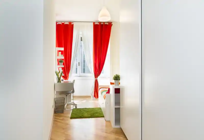 Camera da letto luminosa e moderna con tenda rossa e letto compatto visibile sulla destra. Lo spazio include una scrivania, un tappeto verde e mensole semplici, ideale per un singolo o un annuncio di affitto.
