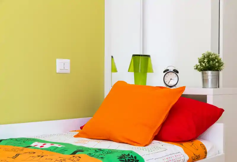 Angolo camera luminoso e colorato con cuscini arancione e rosso su un letto singolo ben rifatto, mensola con sveglia e pianta e parete verde come accent.