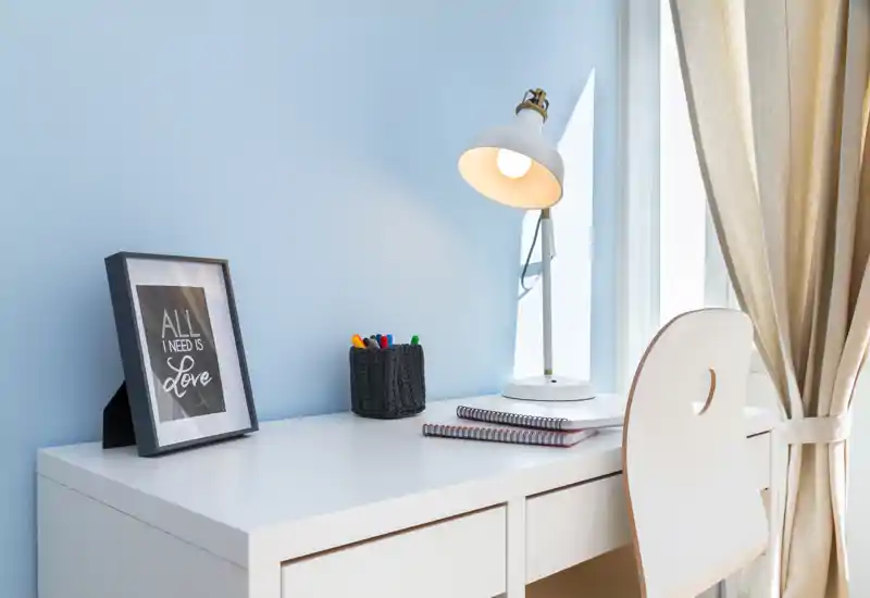 Scrivania minimal e luminosa con lampada moderna, sedia e accessori ordinati vicino a una finestra soleggiata, perfetta per studio o lavoro da remoto.
