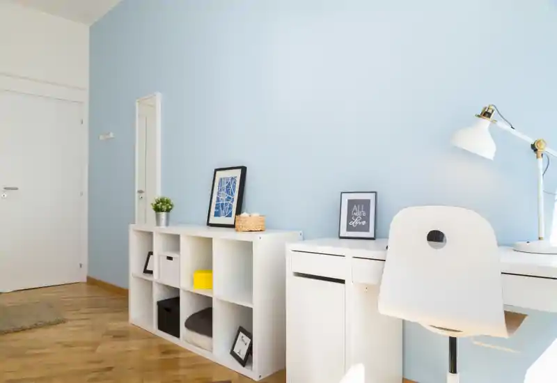 Zona giorno luminosa e moderna con pareti azzurro pallido, scaffalature bianche e una scrivania ordinata. L’ambiente è allestito con stampe incorniciate, tessili morbidi e una lampada da scrivania, ideale per annunci di appartamenti.