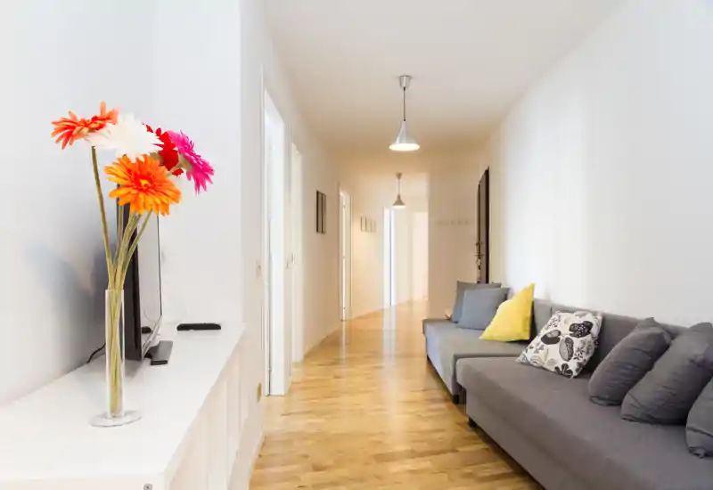 Zona living moderna e luminosa con un lungo corridoio e un divano grigio impreziosito da cuscini colorati e un vaso di fiori; stile minimale e pavimento in legno caldo.