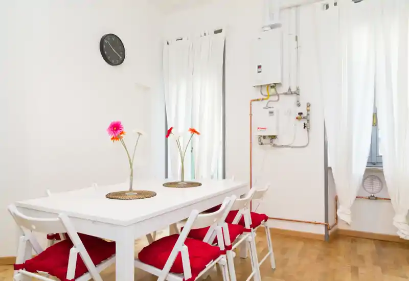 Zona pranzo luminosa e minimal con tavolo bianco, sedie pieghevoli con cuscini rossi e semplici centrotavola floreali. Luce naturale e pavimento in legno chiaro creano un’atmosfera ariosa e accogliente.