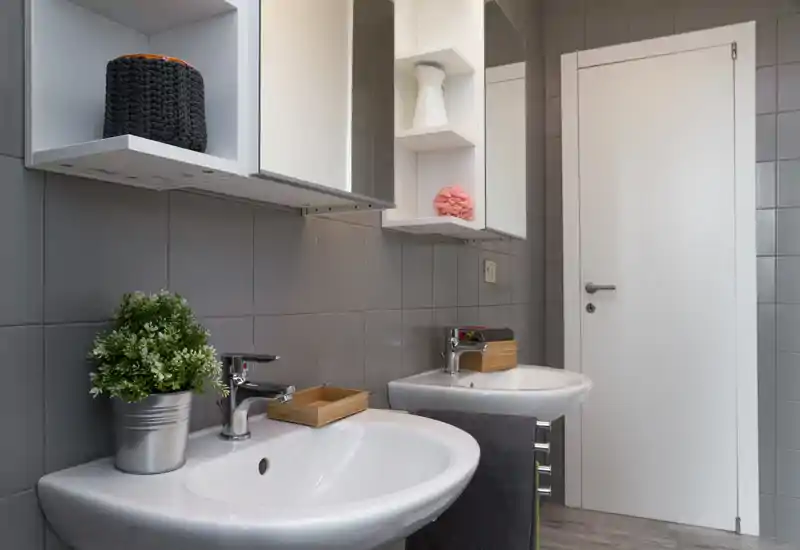 Bagno moderno con due lavabi bianchi, rubinetteria cromata, pareti piastrellate grigie e pensili con specchi — luminoso, pulito e ben allestito.