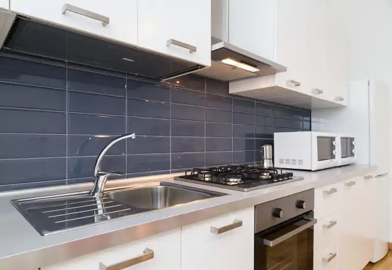 Cucina moderna e luminosa con lavello in acciaio, piano cottura a gas a quattro fuochi e forno integrato. Pensili bianchi e rivestimento scuro per un aspetto pulito e contemporaneo.