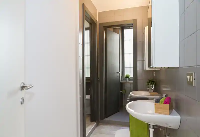 Bagno moderno e pulito con due lavandini a parete, un mobile specchio e piastrelle grigie neutre. La luce naturale entra da una finestra alta sul retro, donando un aspetto fresco e luminoso.