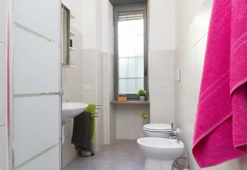 Bagno moderno e luminoso con wc, bidet, lavabo e porta doccia in vetro; piastrelle neutre e una finestra che garantisce luce naturale.