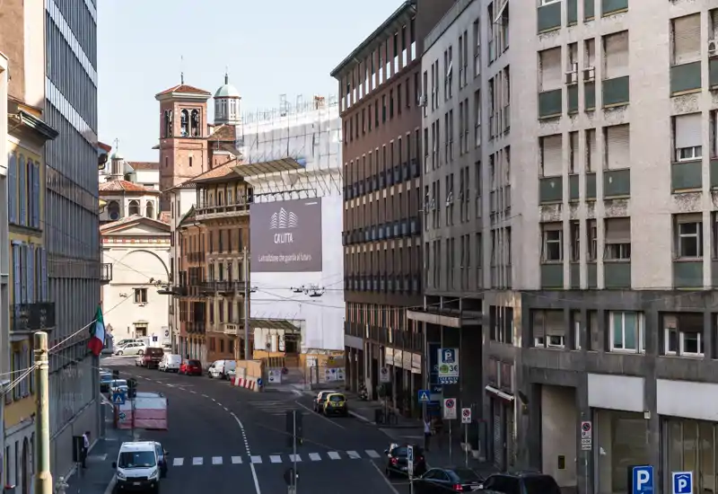 Veduta di una via urbana con palazzi su più piani, auto parcheggiate e una torre campanaria sullo sfondo in pieno giorno — utile per mostrare il quartiere e i collegamenti.