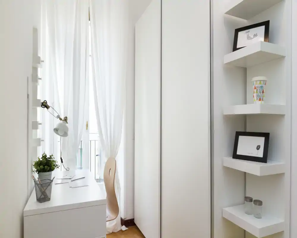 Angolo studio luminoso e minimal con scrivania bianca, sedia in legno e tende leggere che lasciano entrare luce naturale; mensole e piccoli oggetti decorativi rendono il tutto ordinato e accogliente.