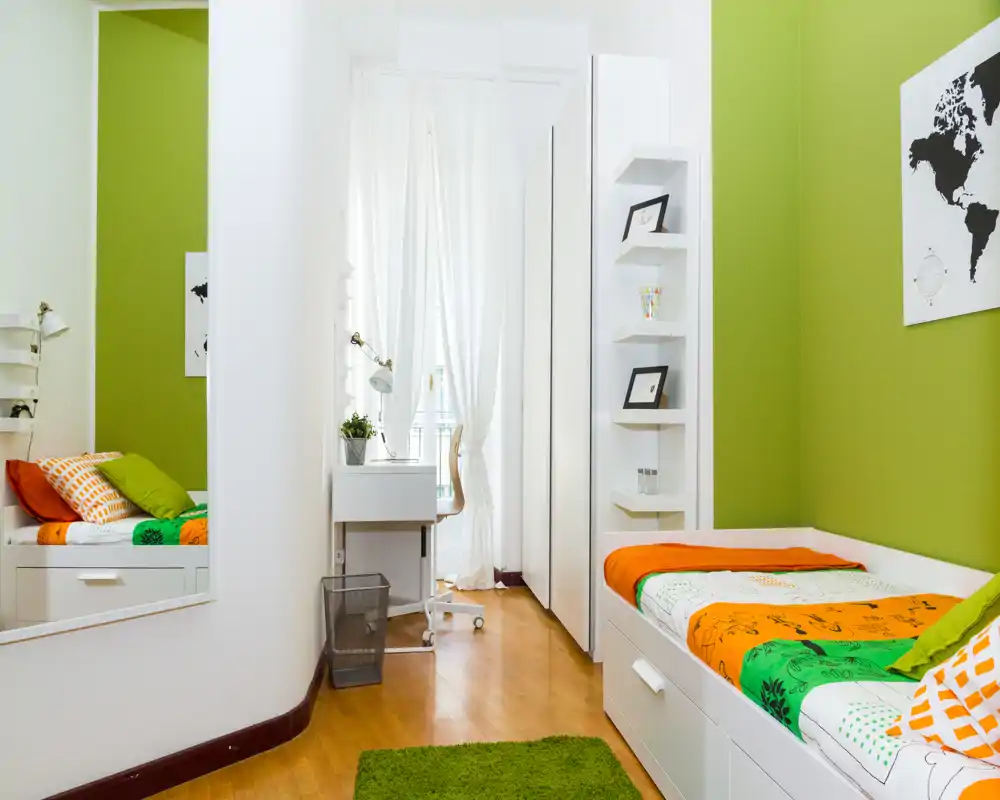Camera singola luminosa e moderna con divano-letto, biancheria colorata e una piccola scrivania vicino alla finestra — perfetta per studenti o affitti brevi.