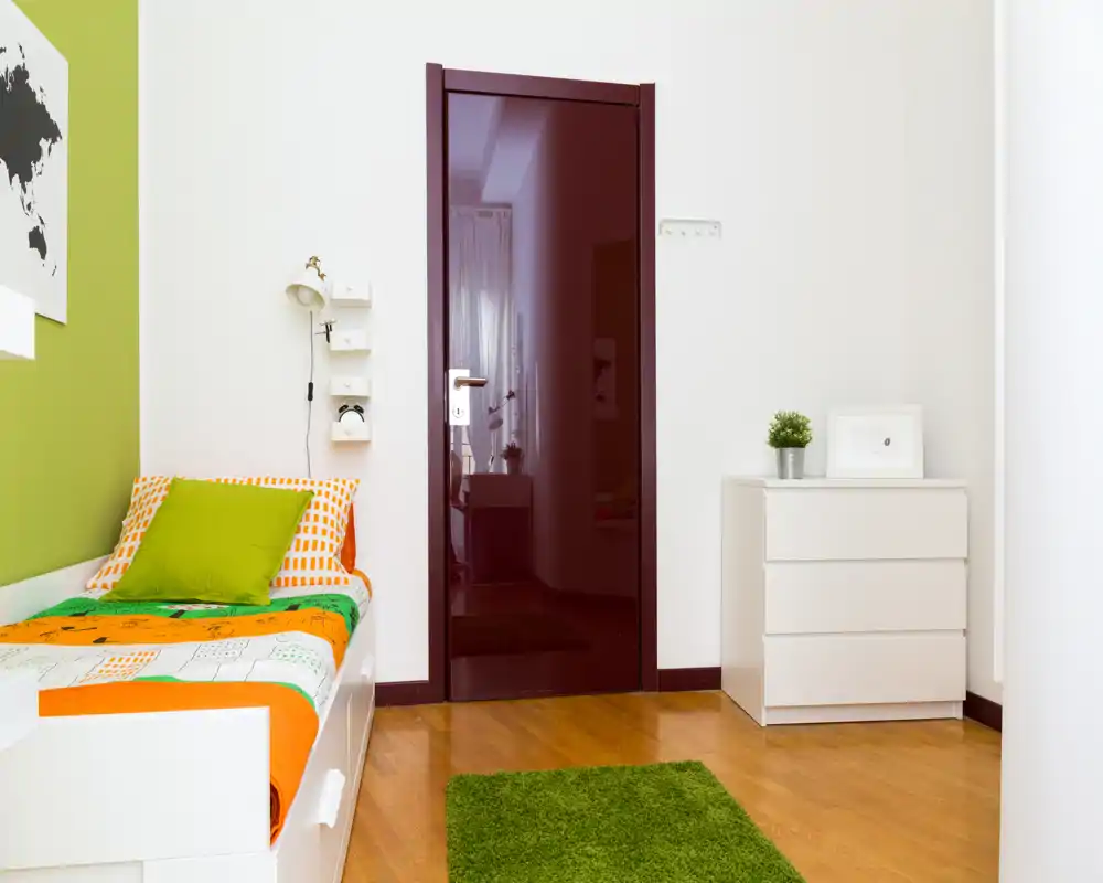 Camera da letto luminosa e compatta con letto singolo con biancheria colorata, un piccolo comò bianco e un tappeto verde che aggiunge calore e contrasto.