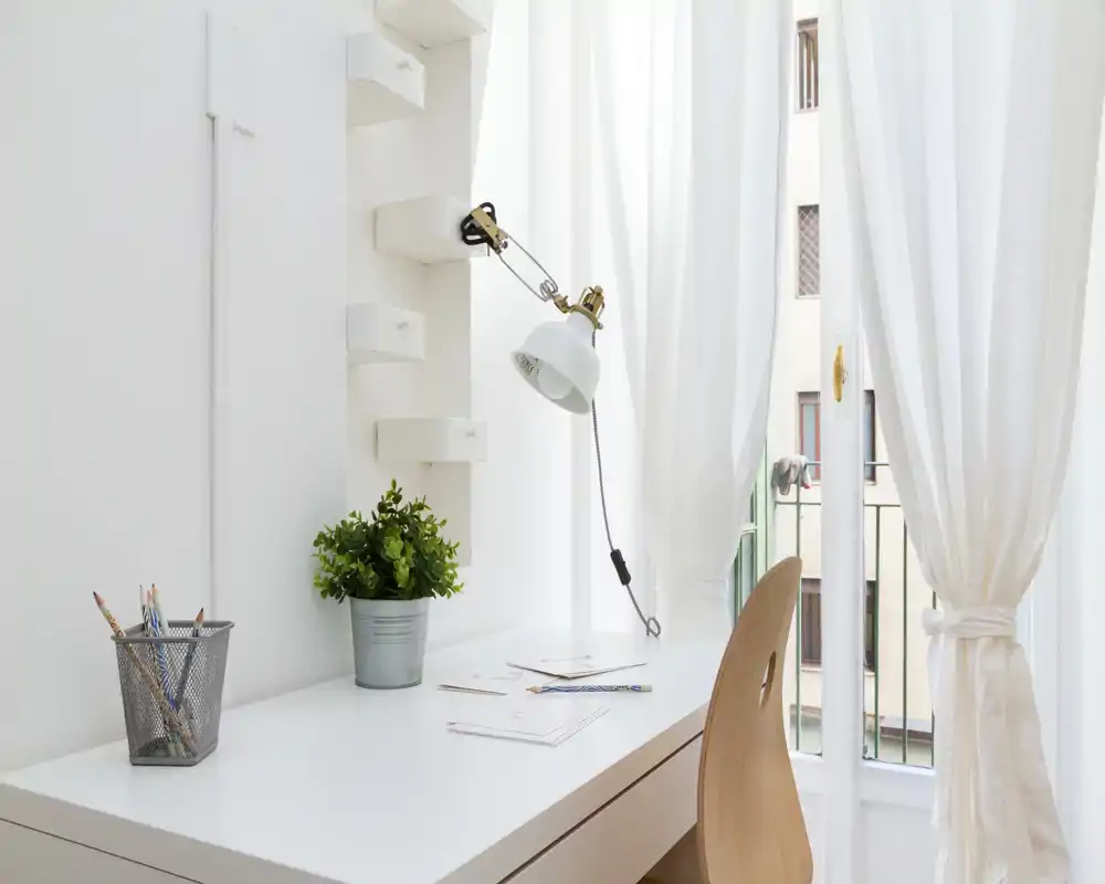 Angolo scrivania luminoso e minimal con sedia in legno, lampada da lavoro e pianta accanto a una finestra soleggiata — perfetto per un piccolo home office o angolo studio.