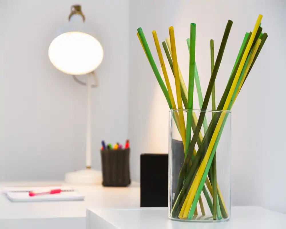 Angolo scrivania luminoso e minimal con un vaso di vetro decorativo con steli verdi e gialli, lampada da tavolo bianca e cancelleria — un dettaglio di workspace moderno e ordinato.